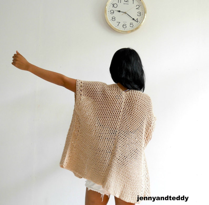 May include: A beige crochet cardigan with a loose, open-weave design. The cardigan is worn by a person with long black hair, standing in front of a white wall with a clock above them.