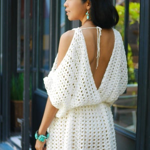 May include: A white crochet dress with a deep V-neck and open shoulders. The dress has a loose fit and a short skirt. The model is wearing turquoise earrings.
