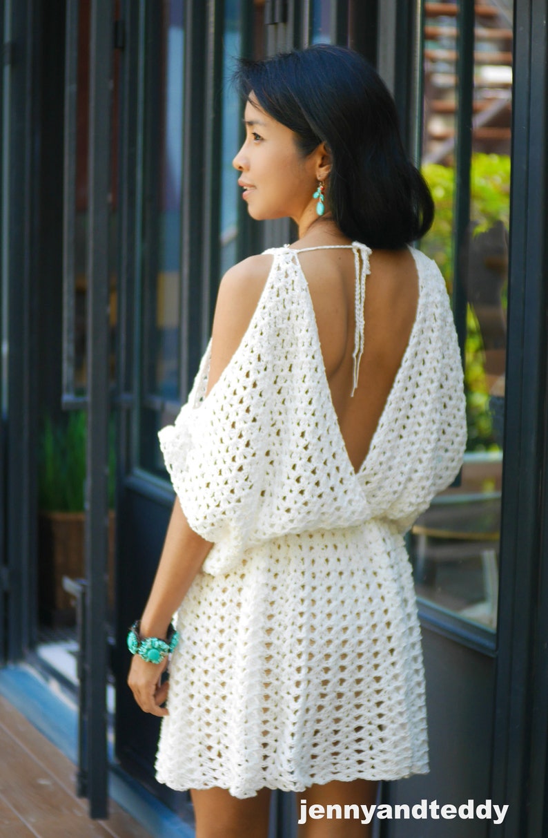 May include: A white crochet dress with a deep V-neck and open shoulders. The dress has a loose fit and a short skirt. The model is wearing turquoise earrings.