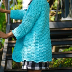 Peut inclure: Un cardigan en crochet turquoise avec un motif de point texturé. Le cardigan est porté sur une jupe à carreaux noirs et blancs.
