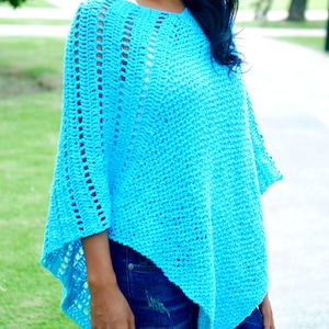 Puede incluir: Un poncho turquesa de ganchillo con diseño de cuello en V. El poncho tiene un ajuste holgado y fluido y presenta detalles calados a lo largo de las mangas. La parte inferior del poncho es asimétrica. El texto "jennyandteddy" es visible en la parte inferior.