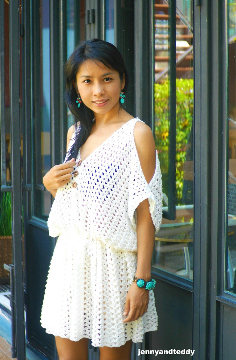 May include: A white crochet dress with a loose fit and a scoop neck. The dress has a crocheted pattern and features a tie at the waist. The model is wearing turquoise earrings and a turquoise bracelet.