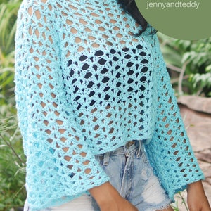 Puede incluir: Un top de crochet azul claro con un patrón de encaje. El top tiene un escote redondo ancho y mangas largas. La modelo lleva el top con pantalones cortos de mezclilla.