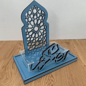 May include: A blue wooden decorative piece with an arched top featuring a geometric pattern. The base has a clear acrylic shelf and a script in black. The piece is designed for display.