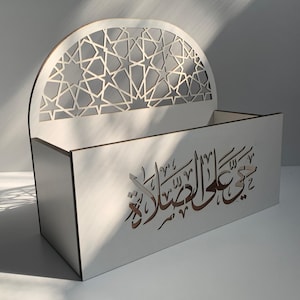 Puede incluir: Caja de madera blanca con parte superior arqueada decorada con un patrón geométrico de estrellas. La parte frontal de la caja muestra caligrafía árabe en color marrón. Diseño moderno con líneas limpias y elementos decorativos.