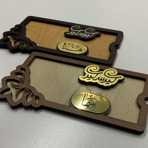 May include: Two wooden plaques with decorative floral corner cutouts. Each plaque has a light-coloured centre, a dark brown frame, and gold-coloured Arabic script. One plaque has the name "Hessa" and the other has the name "Sama".