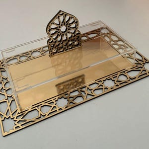 Puede incluir: Una bandeja decorativa con base dorada espejada y tapa de acrílico transparente. La bandeja presenta un diseño geométrico cortado con láser en color marrón oscuro. Un elemento decorativo a juego se encuentra en la parte trasera.