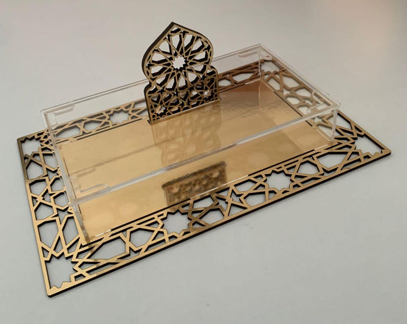 May include: A decorative serving tray with a gold mirrored base and a clear acrylic top. The tray features a laser-cut geometric design in a dark brown color. A matching decorative element stands at the back of the tray.