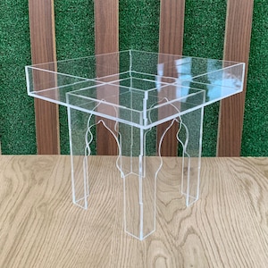 May include: Clear acrylic side table with a square top and decorative arched legs. The table is made of transparent material, offering a modern and minimalist design. The table measures approximately 12 inches in height and 16 inches in width.