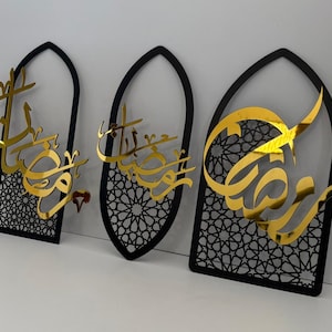 May include: Three decorative wall hangings with a black arched frame and gold Arabic calligraphy. The frames have a geometric pattern in the center. The gold calligraphy is the focal point of the design.