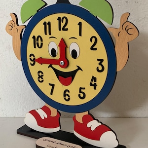 Könnte beinhalten: Eine skurrile Holz-Uhr mit einem lächelnden Gesicht, Händen und Füßen. Das Zifferblatt ist gelb mit schwarzen Zahlen und einem roten Zeiger, der auf 9 zeigt. Die Uhr hat rote Schuhe und grüne glockenförmige Akzente. Ein kleines Holzschild trägt die Aufschrift "Grade 3 B".