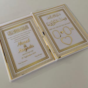 May include: Open white wedding book with gold accents. The book features gold text and decorative borders. The text includes names, a date, and phrases in English and Arabic. The right page has two circles and a heart.