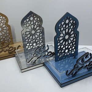 May include: Three decorative wooden trays with intricate arch-shaped cutouts and Arabic script. The trays are in shades of wood, gray, and blue, each with a clear acrylic base. The cutouts feature geometric patterns, and the script is in a flowing, stylized font.