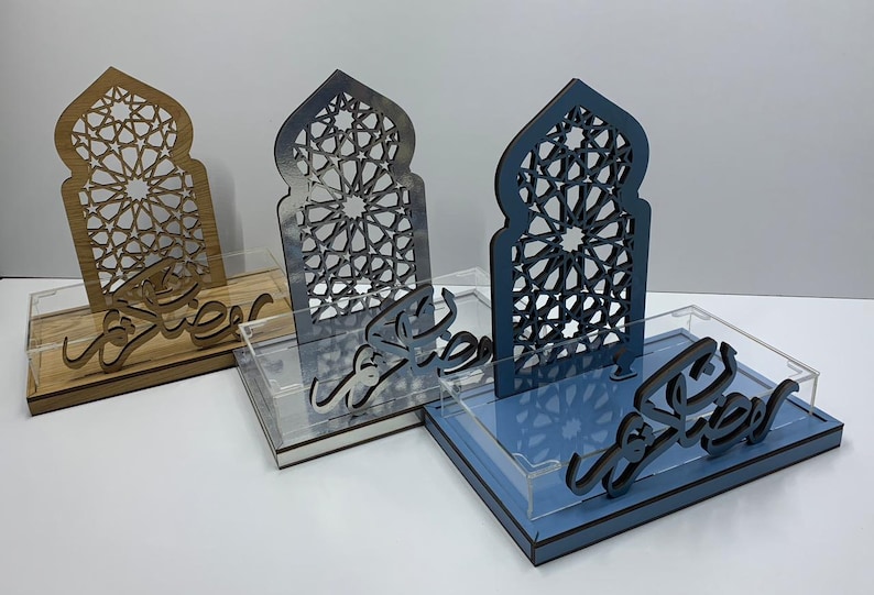 May include: Three decorative wooden trays with intricate arch-shaped cutouts and Arabic script. The trays are in shades of wood, gray, and blue, each with a clear acrylic base. The cutouts feature geometric patterns, and the script is in a flowing, stylized font.