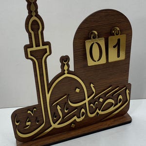 May include: A wooden Ramadan calendar with gold accents. The design includes a mosque silhouette, Arabic calligraphy, and a section for displaying the date. The calendar shows the number "01" in gold.