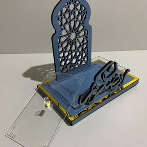 May include: A blue and gold decorative display with Arabic calligraphy. The display features a laser-cut arch design and a clear acrylic cover. The base is blue with gold trim, and the calligraphy is black.