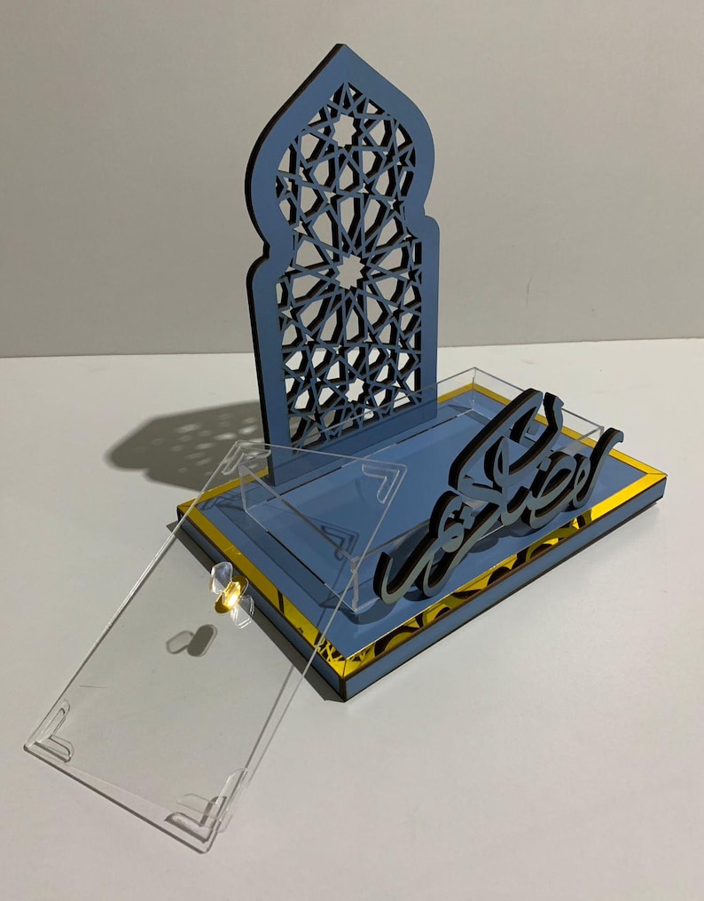 May include: A blue and gold decorative display with Arabic calligraphy. The display features a laser-cut arch design and a clear acrylic cover. The base is blue with gold trim, and the calligraphy is black.