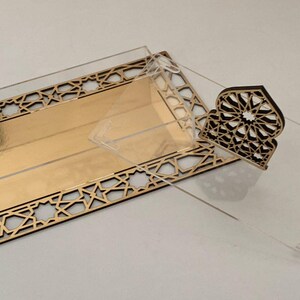 May include: A decorative gold and clear acrylic box with a geometric design. The box has a rectangular shape with a gold interior and a clear lid. A separate decorative piece with a similar design is also included.