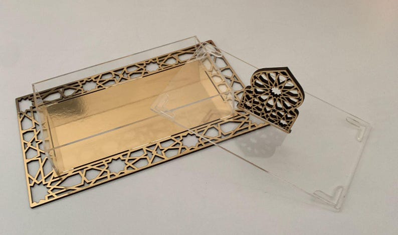 May include: A decorative gold and clear acrylic box with a geometric design. The box has a rectangular shape with a gold interior and a clear lid. A separate decorative piece with a similar design is also included.