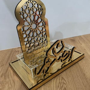 Può includere: Un espositore decorativo dorato con un design geometrico e la parola "Ramadan" in scrittura araba. L'espositore ha una base rettangolare e un vassoio in acrilico trasparente.