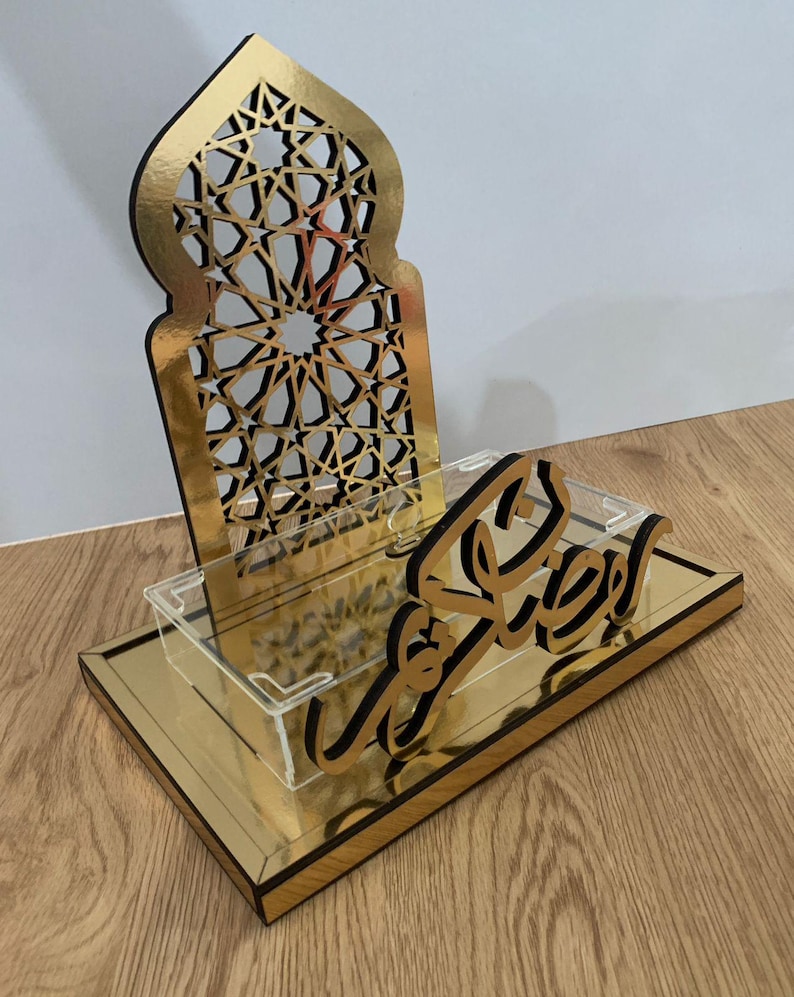 May include: A gold-colored decorative stand with a geometric design and the word "Ramadan" in Arabic script. The stand has a rectangular base and a clear acrylic tray.
