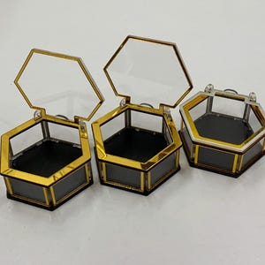 以下が含まれることがあります： ヒンジ付きの透明なガラス蓋が付いた、3つの六角形のディスプレイボックス。ボックスは金色の金属フレームと黒い内装です。ボックスは開いており、内部が明らかになっています。ボックスは白い表面にあります。
