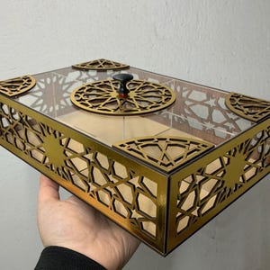 May include: A rectangular wooden box with a clear lid. The box features intricate gold geometric cutouts on the sides and top. A circular gold design with a black knob is on the lid. The box is held by a hand.
