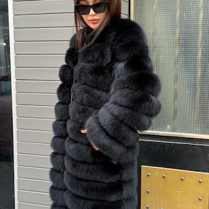 May include: A full-length black fur coat with a layered design. The coat has long sleeves and a plush, textured appearance. The person wearing the coat is also wearing black sunglasses.