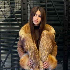 K&ouml;nnte beinhalten: Braune Wildlederjacke mit luxuri&ouml;sem Pelzkragen und Manschetten. Das Fell weist eine Mischung aus goldenen und rotbraunen T&ouml;nen auf, die einen auff&auml;lligen Kontrast zur neutralen Farbe der Jacke bilden. Die Jacke hat einen G&uuml;rtel und wird &uuml;ber einem schwarzen Oberteil getragen.