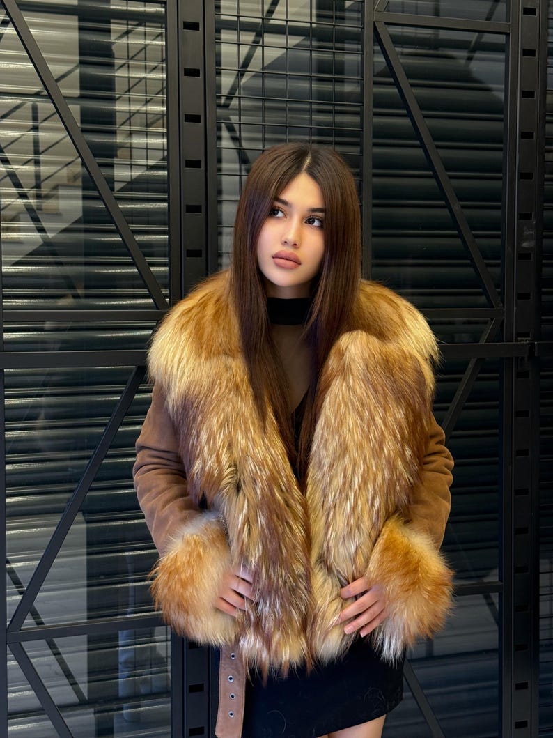 K&ouml;nnte beinhalten: Braune Wildlederjacke mit luxuri&ouml;sem Pelzkragen und Manschetten. Das Fell weist eine Mischung aus goldenen und rotbraunen T&ouml;nen auf, die einen auff&auml;lligen Kontrast zur neutralen Farbe der Jacke bilden. Die Jacke hat einen G&uuml;rtel und wird &uuml;ber einem schwarzen Oberteil getragen.