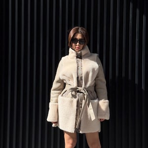 May include: A beige faux fur coat with a high collar, long sleeves, and a tie belt. The coat features a textured design on the front and cuffs, with a patterned trim along the zipper and pockets. The person is wearing black sunglasses and black boots.