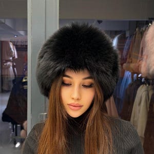 Puede incluir: Un sombrero de piel negro con una corona redondeada. El sombrero está hecho de un material suave y esponjoso. La persona de la imagen lleva un jersey de cuello alto negro.