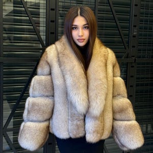 Könnte beinhalten: Ein luxuriöser, hellbrauner Pelzmantel mit breitem Kragen und horizontalen Ärmeldetails. Der Mantel wird über einem schwarzen Oberteil und Kleid getragen, was einen raffinierten und eleganten Look erzeugt. Das Fell wirkt weich und plüschig.