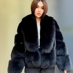 Pode incluir: Casaco de pele preto com gola larga e mangas em camadas. O casaco é feito de um material macio e felpudo e tem uma aparência luxuosa. A modelo está usando um top branco por baixo do casaco.