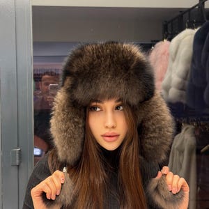 Puede incluir: Un sombrero de piel marrón con orejeras y detalles de pompón de piel a juego. El sombrero es de color marrón oscuro y parece estar hecho de un material suave y lujoso. Un accesorio de invierno.