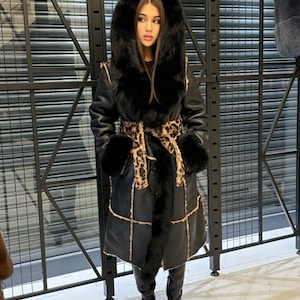 May include: A long black coat with a fur-lined hood and cuffs. The coat has a leopard print belt and trim, and is worn with black boots. The coat appears to be made of leather and fur.