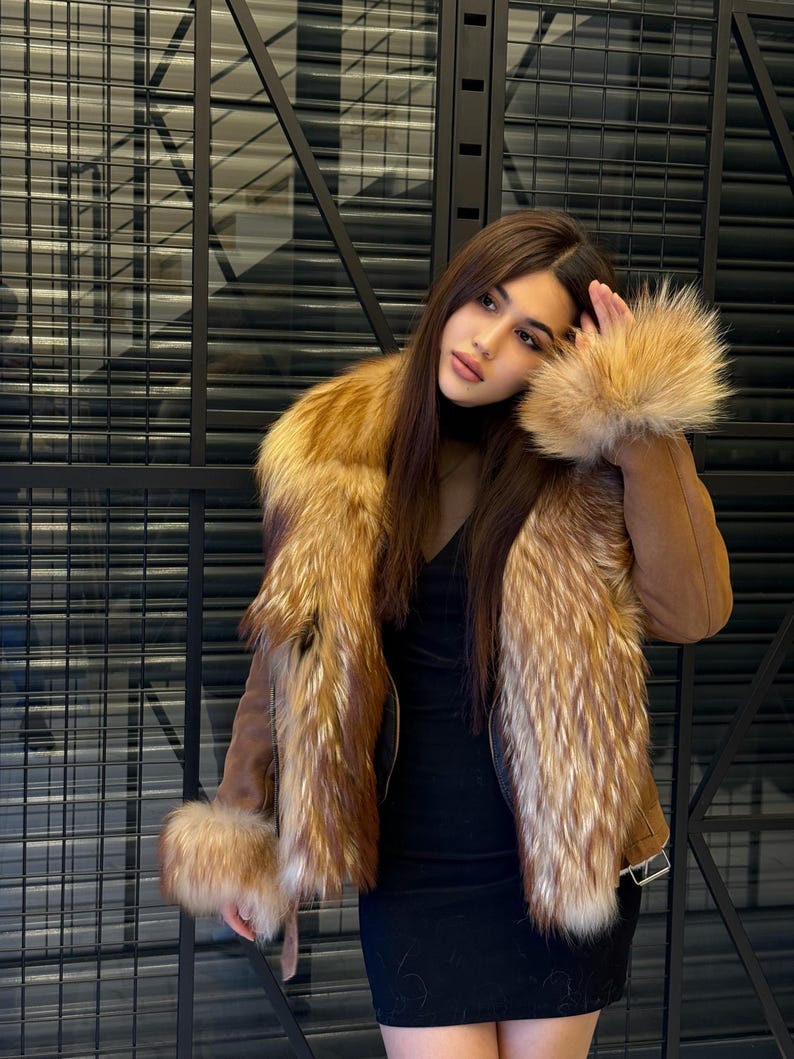 K&ouml;nnte beinhalten: Braune Wildlederjacke mit Pelzkragen und Manschetten. Die Jacke hat einen reichen, goldbraunen Pelz, der mit dem helleren braunen Wildleder kontrastiert. Die Jacke wird &uuml;ber einem schwarzen Kleid getragen.
