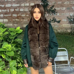 Może przedstawiać: Ciemnozielony płaszcz z brązowym futrzanym kołnierzem i mankietami. Płaszcz jest noszony na czarnym topie. Tło przedstawia ceglany mur, zielone rośliny i metalowe krzesło.