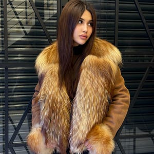 K&ouml;nnte beinhalten: Braune Wildlederjacke mit luxuri&ouml;sem Pelzkragen und Manschetten. Das Fell weist eine Mischung aus goldenen und rotbraunen T&ouml;nen auf, die einen auff&auml;lligen Kontrast zur neutralen Farbe der Jacke bilden. Klassisches Design.