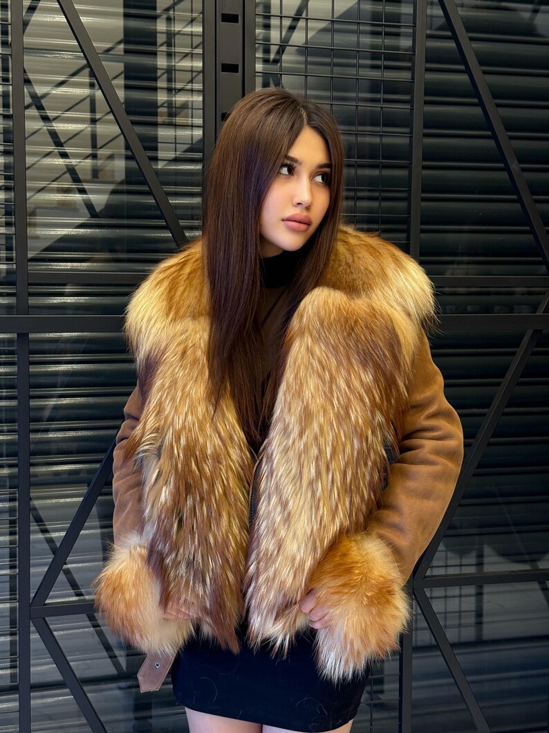 K&ouml;nnte beinhalten: Braune Wildlederjacke mit luxuri&ouml;sem Pelzkragen und Manschetten. Das Fell weist eine Mischung aus goldenen und rotbraunen T&ouml;nen auf, die einen auff&auml;lligen Kontrast zur neutralen Farbe der Jacke bilden. Klassisches Design.