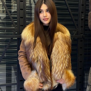 K&ouml;nnte beinhalten: Braune Wildlederjacke mit luxuri&ouml;sem Pelzkragen und Manschetten. Das Fell hat goldene und rotbraune Farbt&ouml;ne, die einen auff&auml;lligen Kontrast zur neutralen Farbe der Jacke bilden. Die Jacke hat einen G&uuml;rtel.