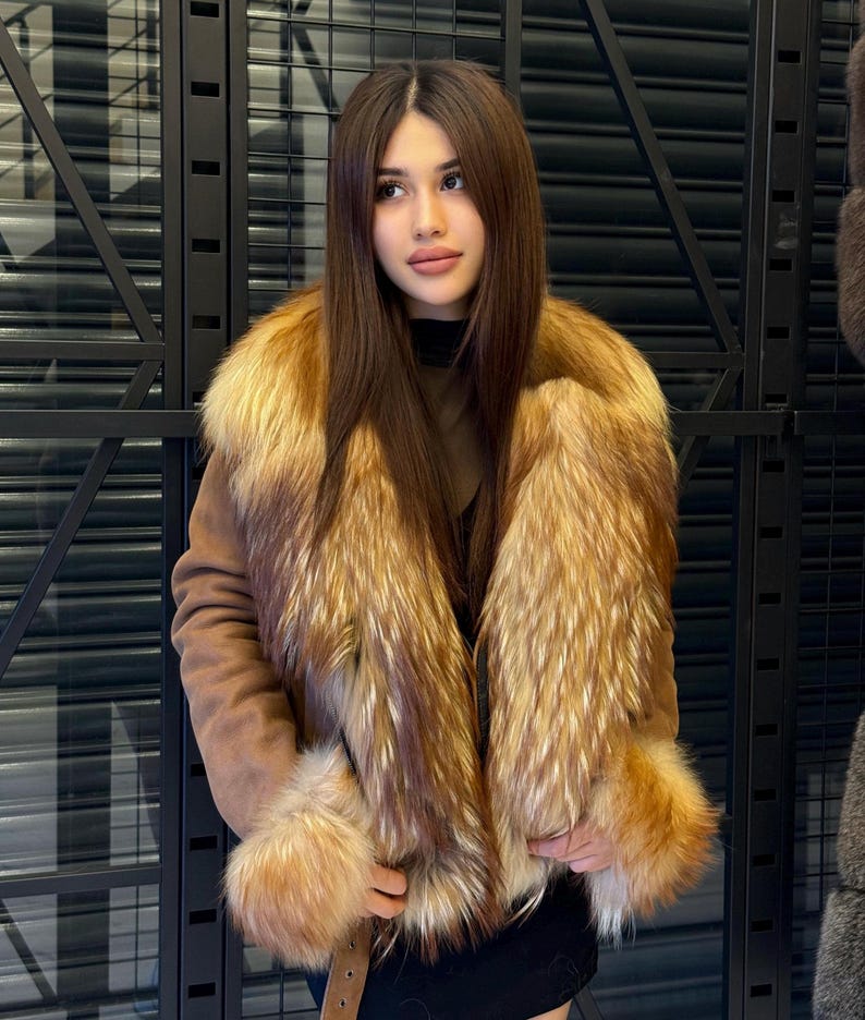 K&ouml;nnte beinhalten: Braune Wildlederjacke mit luxuri&ouml;sem Pelzkragen und Manschetten. Das Fell hat goldene und rotbraune Farbt&ouml;ne, die einen auff&auml;lligen Kontrast zur neutralen Farbe der Jacke bilden. Die Jacke hat einen G&uuml;rtel.