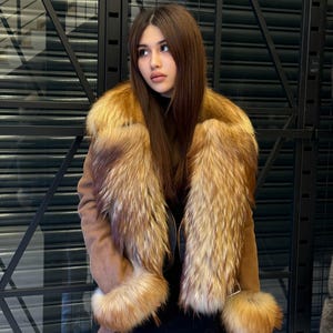 K&ouml;nnte beinhalten: Eine braune Wildlederjacke mit luxuri&ouml;sem Pelzkragen und Manschetten. Die Jacke hat ein dunkles Futter und einen schwarzen Rock. Das Fell ist eine Mischung aus hellen und dunklen Braunt&ouml;nen, die einen reichen, strukturierten Look erzeugt. Die Jacke ist ein stilvolles Oberbekleidungsst&uuml;ck.