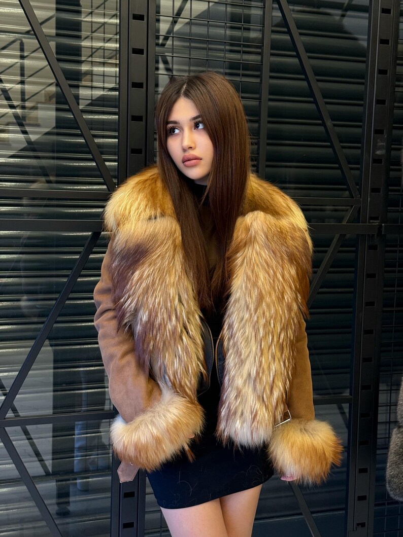 K&ouml;nnte beinhalten: Eine braune Wildlederjacke mit luxuri&ouml;sem Pelzkragen und Manschetten. Die Jacke hat ein dunkles Futter und einen schwarzen Rock. Das Fell ist eine Mischung aus hellen und dunklen Braunt&ouml;nen, die einen reichen, strukturierten Look erzeugt. Die Jacke ist ein stilvolles Oberbekleidungsst&uuml;ck.