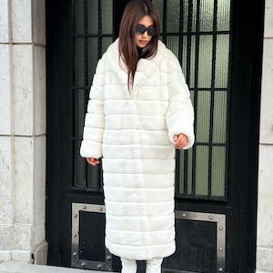 May include: A long, white faux fur coat with a notched collar and a button closure. The coat is floor-length and has a horizontal, ribbed design. The person is wearing white boots and sunglasses.
