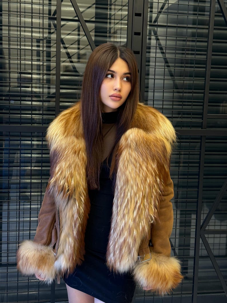 K&ouml;nnte beinhalten: Braune Wildlederjacke mit Pelzkragen und Manschetten. Die Jacke hat einen G&uuml;rtel in der Taille und wird &uuml;ber einem schwarzen Kleid getragen. Das Fell hat helle und dunkle Braunt&ouml;ne. Der Hintergrund ist ein schwarzes Metallgitter.