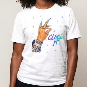 May include: White t-shirt featuring a hand illustration with long, pointed nails, rings, and a watch. The text "CLOCK IT" is written in a gradient of blue and purple. The design is set against a backdrop of small, blue star shapes.