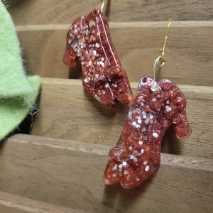 Puede incluir: Un par de pendientes colgantes con un diseño de zapato rojo con purpurina y ganchos dorados.