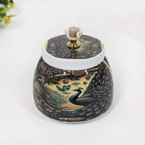 Könnte beinhalten: Ein dekoratives Glas mit Deckel, das ein Pfauenmuster aufweist. Das Glas hat einen weißen Rand und einen durchsichtigen Kristallknopf. Der Pfau ist mit ausgebreiteten Federn dargestellt, und das Glas hat eine detaillierte, farbenfrohe Illustration.