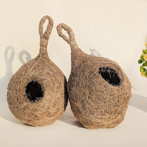 Könnte beinhalten: Zwei handgefertigte, braun gewebte Vogelhäuser mit kreisförmigen Eingangsöffnungen und geschlungenen Aufhängegriffen. Die Vogelhäuser sind aus Naturfasern gefertigt und haben ein rustikales Aussehen. Der Hintergrund ist eine schlichte weiße Oberfläche.